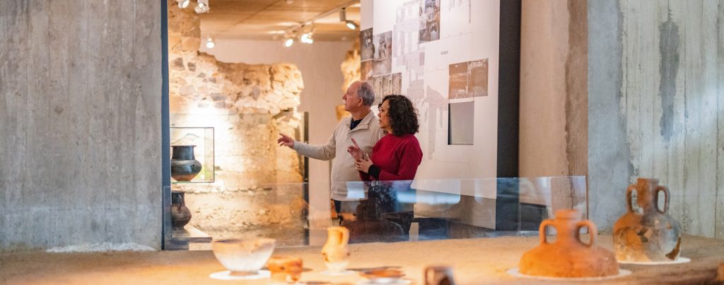 Two people in a museum setting looking at a display on a wall, with ancient earthenware in the foreground