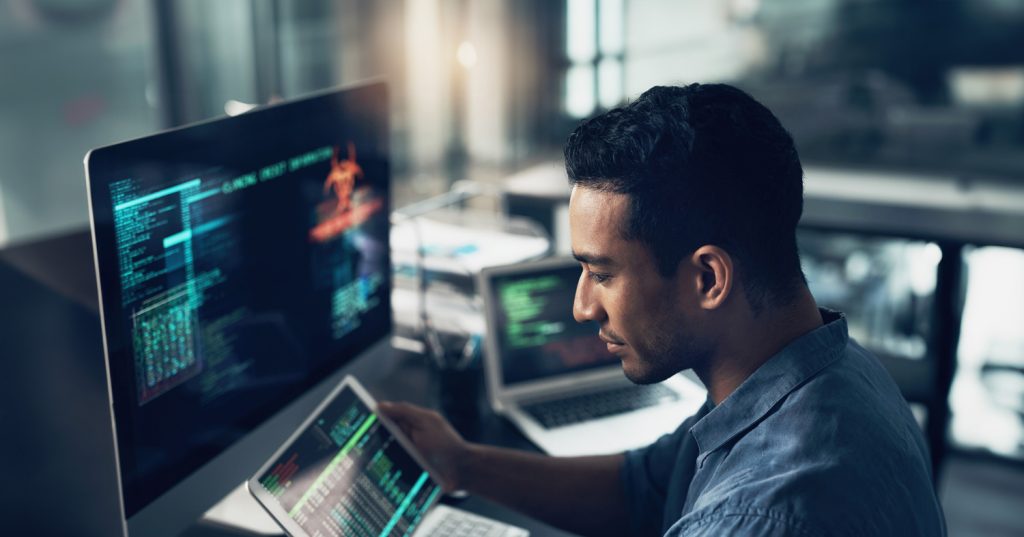 Man in an office setting looks at a tablet screen with lines of code, with a laptop and computer monitor in the background