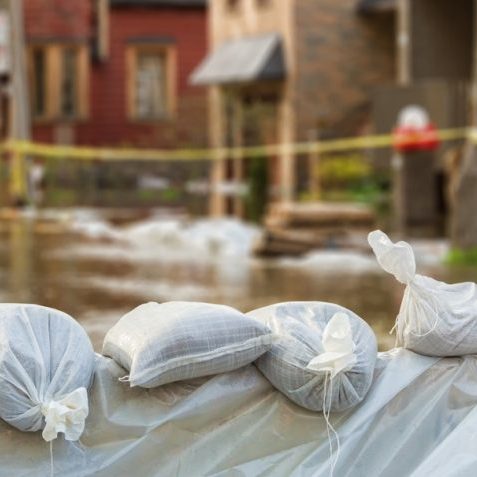 A pile of sandbags in the foreground and a group of houses with caution tape in the background