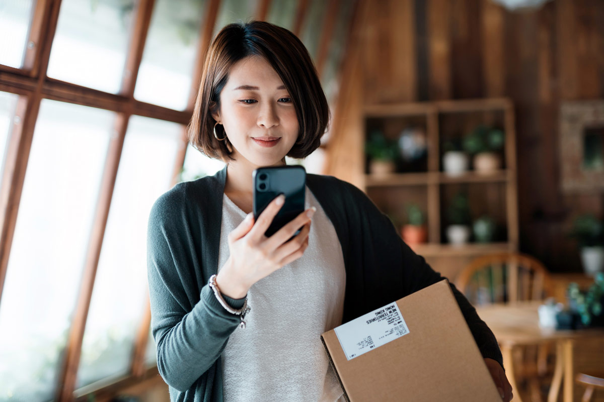 Woman with a box packaged to ship looks at her phone smiling