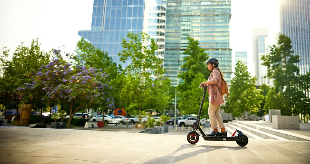 Person riding an electric scooter along a city sidewalk, with a city skyline in the background on a sunny day