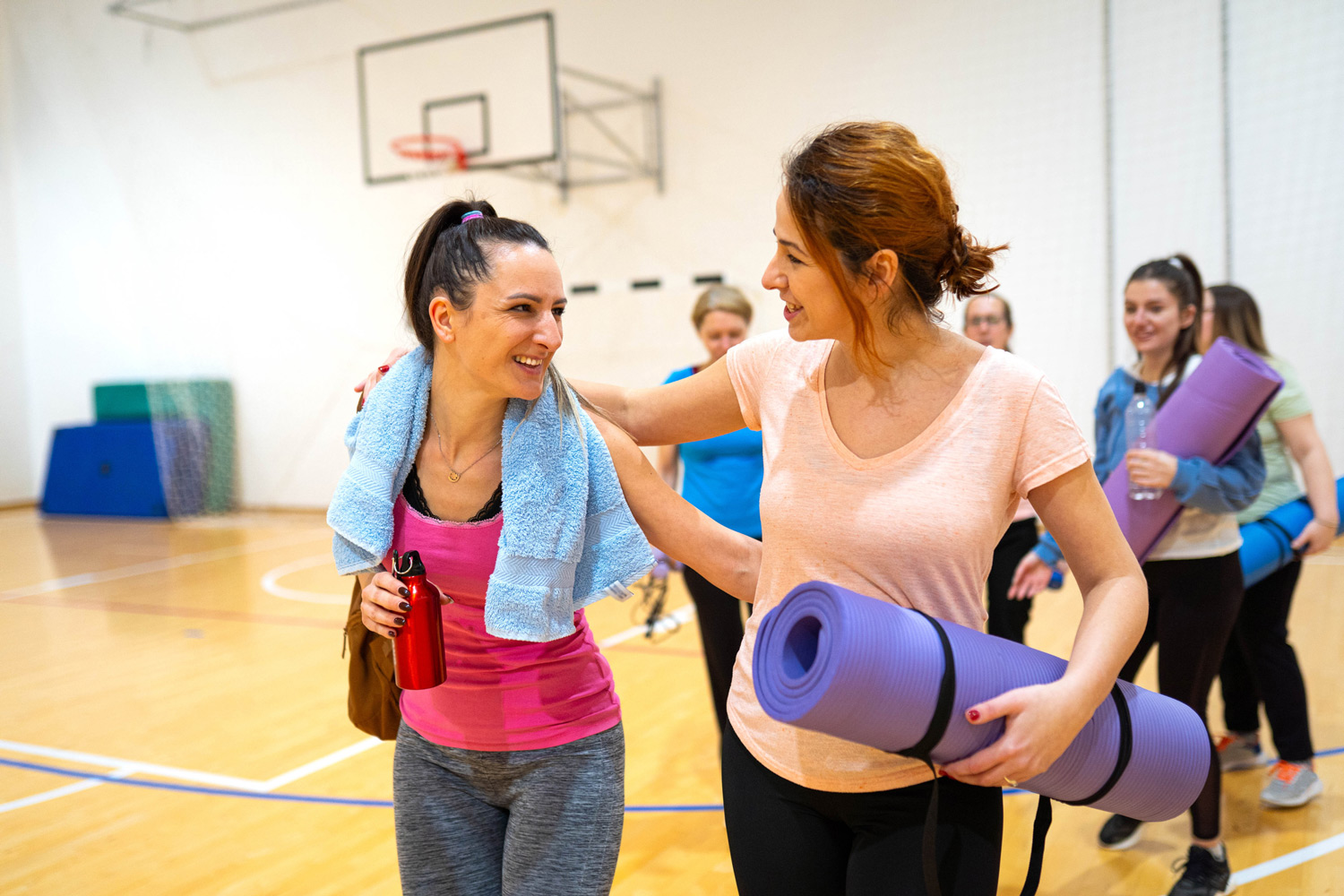 Two women with yoga mats have a chat after their class. Gymnasium setting in the background.