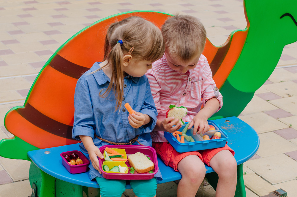 Two little kids sit and eat healthy snacks out of their lunchboxes