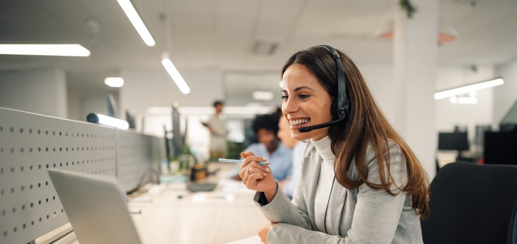 Young woman uses a headset and points to her computer screen, in a brightly lit office