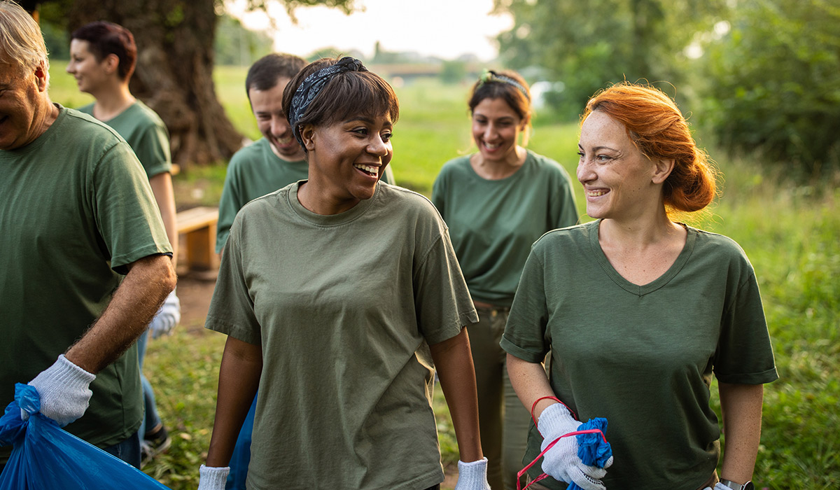 Two volunteers in matching shirts smile at each other as they walk through a greenspace with recycling bags